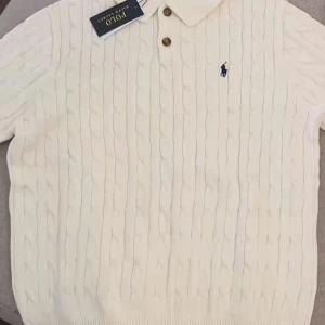 Vit kabelstickad pikétröja Polo Ralph Lauren - Kabelstickad vit pikétröja från Polo Ralph Lauren med klassisk krage, två bruna knappar och broderad blå logga på bröstet. Tröjan har lång ärm och är gjord i mjuk bomull. Perfekt för en clean och stilren look.