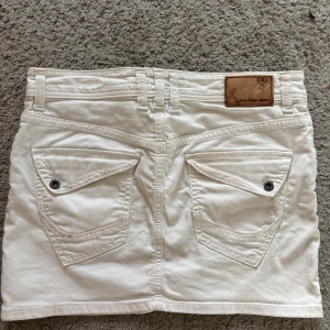 Vit jeanskjol från Calvin Klein Jeans - Snygg vit kort jeanskjol från Calvin Klein Jeans med klassiska fickor både fram och bak, bälteshällor och knapp- samt dragkedjestängning. Gjord i bomull med lite stretch för skön passform. Perfekt till sneakers eller boots för en cool look.