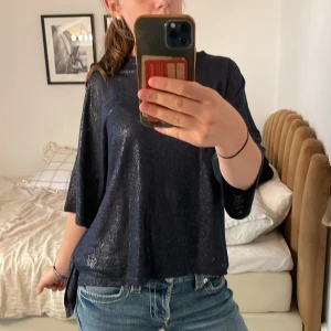 Glittrig mörkblå topp med broderi - Säljer en glittrig mörkblå topp med lös passform och trekvartsärm. Tyget har en skimrande finish och ett litet broderat motiv i silver vid nederkanten. Perfekt för dig som vill sticka ut lite extra med en cool och bekväm stil.