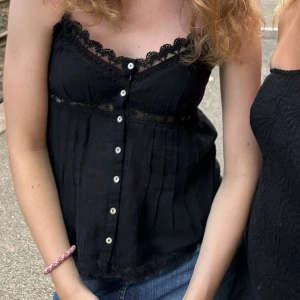Svart spetslinnetopp med knappar - Supersöt svart linnetopp med spetsdetaljer längs urringningen och under bysten. Toppen har knappar framtill och tunna axelband. Perfekt för en chill och snygg look till jeans eller kjol.