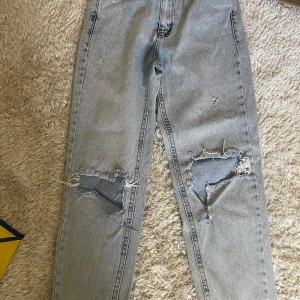Ljusgrå ripped jeans med raka ben - Säljer ett par ljusgrå jeans med raka ben och stora slitningar över knäna. Jeansen har klassisk femficksdesign och hög midja. Perfekta för dig som gillar en avslappnad och edgy stil.