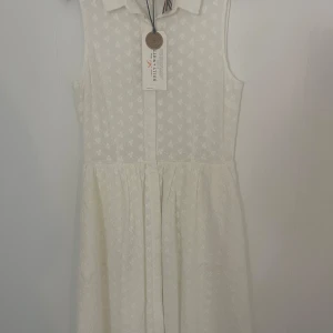 Vit blommig klänning Holly & Whyte - Superfin vit klänning från Holly & Whyte med diskret blommigt mönster, klassisk krage och knäppning framtill. Ärmlös , perfekt för en fräsch och somrig look. Tillverkad i bomull för en luftig känsla.