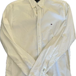 Tommy Hilfiger skjorta  - Klassisk vit skjorta från Tommy Hilfiger – stilren, tidlös och alltid rätt. Skjortan har en snygg passform och diskret logobroderi vid bröstet, vilket gör den perfekt både till vardags, jobb eller mer uppklädda tillfällen. Ett måste i varje garderob! Ren, fräsch och utan defekter – redo att användas!