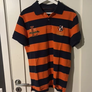 Randig orange och marinblå pikétröja - Snygg pikétröja från Lexington med breda orange och marinblå ränder, klassisk krage och broderade detaljer på bröstet. Kortärmad modell i bomull som ger en sportig och preppy vibe. Perfekt för dig som gillar färg och stil med attityd.