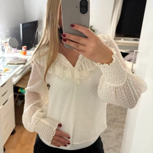 Vit blus med volang och prickigt mönster - Supersöt vit blus från Zara med diskret prickigt mönster, volangdetaljer vid halsen och breda smockade ärmslut. Lätt transparent material, perfekt för dig som gillar en feminin och trendig stil.