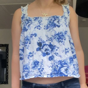 Blommig vit och blå linnetopp - Supersöt linnetopp med breda axelband. Toppens vita tyg är täckt av blåa blommor som ger en fräsch och somrig vibe. Perfekt till jeans eller kjol för en chill look. Men passar även till hösten och vinter med en fin kofta.  Hör av dig vid några frågor💓