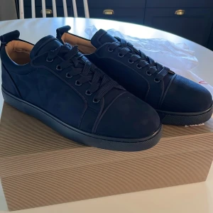 Mörkblå sneakers från Louboutin - !!Priset kan diskuteras vid snabbaffär!!Snygga mörkblå sneakers från Louboutin i mocka med klassisk röd sula och broderad logga på hälen. Skorna har rund tå, platt sula och snörning. Insidan är fodrad i beige läder för extra komfort. Perfekta för dig som vill sticka ut med exklusiv stil. 