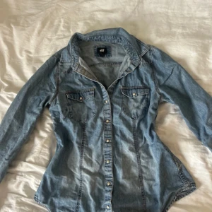 Figursydd jeansskjorta från H&M - Snygg jeansskjorta från H&M med klassisk krage, tryckknappar och två bröstfickor. Skjortan har lång ärm och är figursydd för en schysst passform. Perfekt nu till hösten och i utmärkt skick!
