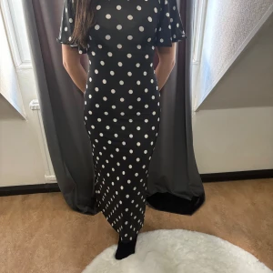 Svart prickig långklänning - Supersnygg svart långklänning med vita prickar och korta, vida ärmar. Klänningen har en figurnära passform som framhäver siluetten och ett klassiskt prickigt mönster som ger retrovibbar. Perfekt för dig som vill sticka ut med en tidlös look.