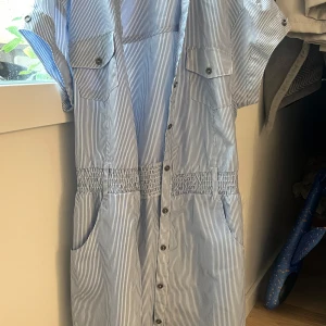 Blåvit randig skjortklänning från Mood  - Snygg blåvit randig skjortklänning med korta ärmar, knappar hela vägen framtill och två bröstfickor med lock. Klänningen har resår i midjan för en skön passform och praktiska sidfickor. Perfekt för en chill sommardag.