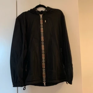 Svart vindjacka från Burberry - Svart vindjacka från Burberry med klassiskt rutigt mönster längs dragkedjan och i huvan. Jackan har huva med snörning, dragkedja framtill och meshfoder på insidan. Perfekt för dig som vill ha en stilren och ikonisk look.