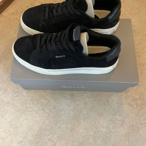 Svarta mockasneakers från Gant - Stilrena svarta sneakers från Gant i mocka med vit platt sula och diskret logga på sidan. Skorna har snörning och en klassisk rund tå. Perfekta för dig som gillar en clean och tidlös look.