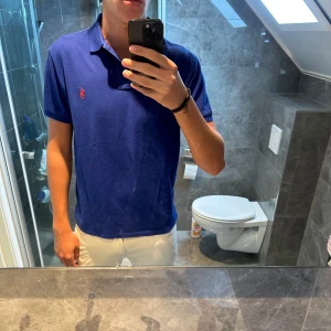 Blå pikétröja från Polo Ralph Lauren - Säljer en klassisk blå pikétröja från Polo Ralph Lauren i modellen Custom Slim Fit. Tröjan har korta ärmar, krage och den ikoniska röda loggan broderad på bröstet. Tillverkad i mjuk bomull som är skön att bära hela dagen.