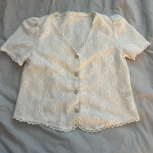 Vit blus med pärlknappar Shein - Superfin vit blus från Shein i storlek S, med broderade spetsdetaljer och v-ringning. Korta puffärmar och pärlknappar framtill. Blusen har vågig spetskant längs ärmar och nederkant.