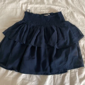 Mörkblå volangkjol från The New - Supersöt mörkblå kortkjol från The New med bred smock i midjan och dubbla lager volanger. Kjolen har en flowy känsla och passar perfekt till sneakers eller sandaler. Materialet känns lätt och luftigt, perfekt för varmare dagar.