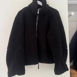 Svart teddyjacka från H&M, storlek M - Mysig svart teddyjacka från H&M i storlek M. Jackan har hög krage, dragkedja framtill och en oversized passform. Perfekt för kalla dagar när du vill vara både varm och stilren. Materialet är fluffigt och ger en cool look.