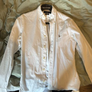 Vit slim fit skjorta från Ralph Lauren - Klassisk vit skjorta från Ralph Lauren i slim fit-modell. Skjortan är tillverkad i stretchig bomull, har långa ärmar, knappar framtill och den ikoniska broderade loggan på bröstet. Perfekt för en clean och stilren look.