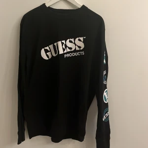 Svart långärmad tröja från Guess - Svart långärmad tröja från Guess med stor vit logga på bröstet och coola tryck längs ena ärmen i vitt och turkost. Tröjan har en klassisk rund halsringning och är tillverkad i mjuk bomull, perfekt för en avslappnad streetstyle-look.