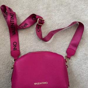Rosa axelväska från Valentino - Säljer en snygg axelväska från Valentino i stark rosa färg med justerbart axelband där loggan står tryckt. Väskan är i präglat skinnliknande material, har guldfärgad dragkedja och ett rymligt fack med innerficka. Perfekt för dig som vill sticka ut med en färgklick!