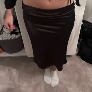 Svart satinkjol i midilängd - Säljer en svart, glansig satinkjol med midilängd. Kjolen har en slät och figurnära passform som faller snyggt längs kroppen. Perfekt för dig som gillar en stilren och elegant look. Passar till crop tops eller stickade tröjor.