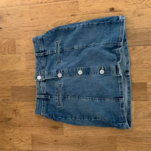 Jeanskjol med knappar från Lindex - Snygg jeanskjol från Lindex med raka linjer och fyra silvriga knappar framtill. Kjolen har bälteshällor och klassisk blå denimfärg. Kan även användas som en tub topp. 