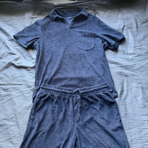 Mörkblå frottéshorts och piké Race Marine - Mörkblå shorts och piké från Race Marine i mjuk frotté med elastisk midja och snörning. Avslappnad passform, perfekt för chill dagar eller strandhäng. Shortsen har fickor på sidorna och en bakficka. Pikén har en bröst ficka på vänstersidan av tröjan. Skandinavisk sportig stil och riktigt sköna att bära. Piken i storlek S och shortsen i storlek M