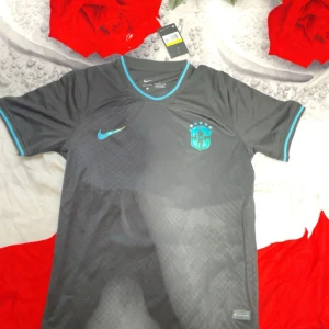 Svart Brasilien Nike fotbollströja S - Snygg svart Brasilien fotbollströja från Nike med blå detaljer och CBF-emblem på bröstet. Tillverkad i Dri-FIT-material som andas, perfekt för träning eller match. Kortärmad modell med rund hals och blå kantband vid ärmslut och krage.