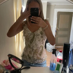 Blommig linnetopp från Saint Tropez - Supersöt linnetopp från Saint Tropez i vitt med blommigt mönster i grönt och beige. Toppen har volangiga axelband, v-ringning och lätt figursydd passform. Perfekt för varma dagar och ger en romantisk vibe till din outfit. Det är en M men passar även S