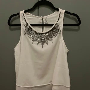 Vit topp med paljettdetaljer - Säljer en vit, ärmlös linne-topp från stradivarius aldrig använt!!  Toppen har en flowy passform, paljett detaljer, och knäpps med en knapp i nacken. 