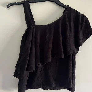 Svart oneshoulder topp från Ellos - Säljer en svart oneshoulder topp från Ellos i mjuk bomull. Toppen har volangdetalj över axeln och snörning i sidan som ger en cool look. Perfekt för dig som vill sticka ut lite extra med en asymmetrisk stil.