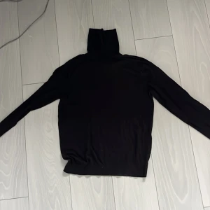 Svart polotröja från Benetton - Klassisk svart polotröja från United Colors of Benetton. Tröjan har lång ärm och hög polokrage, perfekt för lager-på-lager. Mjuk och tunn i materialet, passar snyggt till både jeans och kjol. Enkel och stilren design som funkar året om.