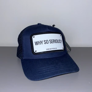Blå keps John Hatter & Co Why So Serious - Mörkblå truckerkeps från John Hatter & Co med vit skylt framtill där det står 'WHY SO SERIOUS?'. Kepsen har böjd skärm, mesh-bakstycke och justerbar snapback. Snygg och modern accessoar med cool attityd.
