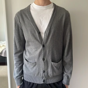 Grå kofta från Blueprint, storlek M - Snygg grå kofta från Blueprint i storlek M. Modellen har v-ringning, knäppning framtill med fyra knappar och två fickor. Materialet är mjukt och tunt, perfekt för lager-på-lager. Passar dig som gillar en clean och stilren look.