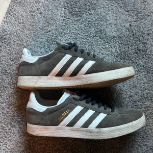 Adidas Gazelle grå mockasneakers - Säljer ett par klassiska Adidas Gazelle sneakers i grå mocka med vita detaljer och svarta snören. Skorna har inte används mycket! Skorna är 39 1/3 och passar både killar och tjejer! Säljer pga att de inte kommer till användning och för att de är för små skorna är i väldigt bra skick är lite smutsiga men kan tvätta innan jag fraktar! Skriv om ni undrar nåt! MVH Arvid 