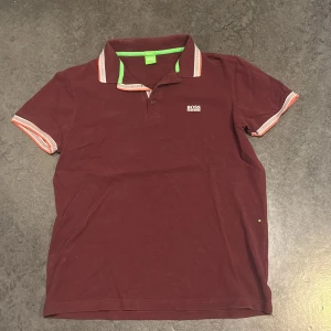 Vinröd kortärmad piké från BOSS - Vinröd pikétröja från BOSS med vita och orange detaljer på krage och ärmslut. Klassisk polo med knappslå och liten BOSS-logga på bröstet. Skön bomullskvalitet och snygg passform, perfekt till jeans eller shorts.