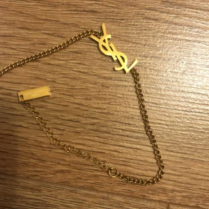 Guldigt YSL halsband från beauticoo  - Snyggt halsband i guldtonad metall från Saint Laurent med den ikoniska YSL-loggan framtill. Kedjan är tunn och elegant med en liten bricka där det står 'Saint Laurent Paris'. Perfekt för dig som vill ha en lyxig och trendig accessoar.