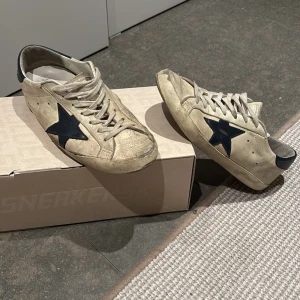 Beige Golden Goose sneakers med svart stjärna - Golden Goose sneakers i beige skinn med ikonisk blå stjärna på sidan och blå hälparti med logga. Klassisk låg modell med snörning och platt sula. Snyggt slitna detaljer för en cool, avslappnad vibe. Skorna levereras med originalkartong och dustbag.