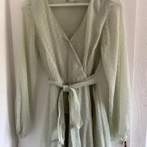 Ljusgrön omlottklänning med bälte - Superfin ljusgrön omlottklänning från Nelly med knytbälte i midjan och långärmade, luftiga ärmar. Klänningen har diskret prickigt mönster och v-ringad hals. Perfekt för vår och sommar!