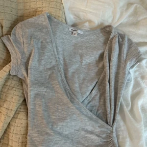 Grå omlottopp från Subdued XS - Snygg ljusgrå omlottopp från Subdued i mjuk bomull. Toppen har korta ärmar, v-ringning och lätt rynk vid sidan för en schysst passform. Perfekt att styla med jeans eller kjol för en chill look.