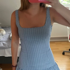 Blå- och vitrandig kort klänning PLT - Supersöt blå- och vitrandig kort klänning från PrettyLittleThing med fyrkantig urringning och breda axelband. Klänningen har en figurnära passform upptill och volang nertill, samt dragkedja i ryggen. Perfekt för sommarens alla tillfällen!