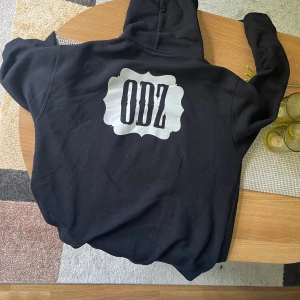 Svart ODZ hoodie med tryck - Svart hoodie från ODZ med stort vitt tryck på ryggen och mindre logga på bröstet. Klassisk känguruficka framtill, huva med snörning och långa ärmar. Perfekt för dig som gillar streetwear och vill sticka ut med en fet logga.