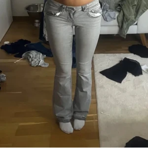 Grå bootcut jeans med fickdetaljer - Snygga ljusgrå jeans med bootcut passform och coola fickor med lock och knapp både fram och bak. Jeansen har klassisk femficksdesign och markerade sömmar. Perfekta för dig som gillar en retrovibe och vill sticka ut lite extra.