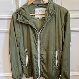 Olivgrön vindjacka från Woolrich - Snygg olivgrön vindjacka från Woolrich med huva och vita dragkedjor framtill och på fickorna. Jackan är lätt och sitter bra. Materialet är tunt och smidigt.;