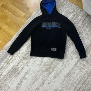 Hoodrich hoodie  - Svart hoodie från Hoodrich med blå text och detaljer på bröstet och ryggen. Insidan av huvan är blå, och tröjan har snörning i huvan samt en klassisk känguruficka framtill. Tröjan är nyskick, storlek M.