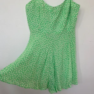 Grön blommig jumpsuit - Superfin grön kortklänning med små vita blommor och smala axelband. Jumpsuiten/ byxdressen har en lätt utsvängd kjol och v-ringad halsringning. Använd 1 gång.