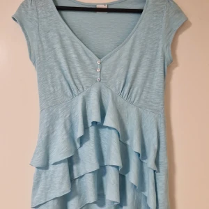 Ljusblå volangblus från Gina Tricot - Supersöt ljusblå blus med v-ringning och tre små knappar framtill. Toppen har korta ärmar och flera lager volanger framtill som ger en flowy känsla. Perfekt för dig som gillar romantiska detaljer och en luftig stil.
