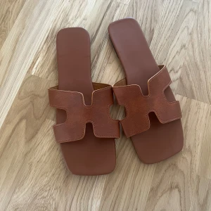 Bruna sandaler  - Nya och oanvända snygga bruna sandaler med breda remmar i skin. Perfekta för varma dagar och ger en stilren look till din outfit. Slip-on modell med platt sula och minimalistisk design.