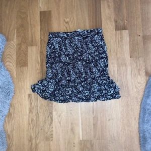 Svart blommig volangkjol från H&M - Supersöt svart kortkjol från H&M med vita små blommor och volang nertill. Kjolen har en bekväm resår i midjan och är gjord i lätt polyester, perfekt för en chill och trendig look.