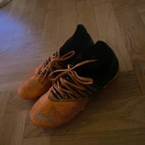 Orangea och svarta fotbollsskor från Puma - Snygga fotbollsskor från Puma i orange och svart med coola mönster och snörning. Ovandelen har textur för bättre bollkontroll och skon har en strumpliknande passform runt ankeln. de är lite smutsiga på ovansidan, men de tvättas självklart innan jag postar. Perfekta för dig som vill sticka ut på planen.