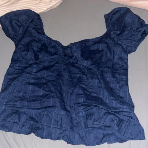 Mörkblå blus med volang från Nelly - Supersöt mörkblå blus från Nelly med korta puffärmar, knappar framtill och volangdetalj längst ner. Blusen har en lätt struktur i tyget och en feminin, lite romantisk look. Perfekt att styla med jeans eller kjol för en trendig vibe.